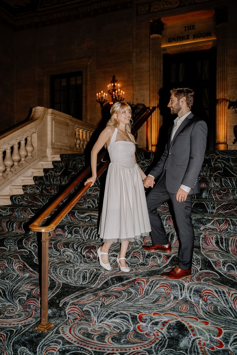 Couple walking on ornate patterned carpet in an elegant hallway with marble stairs and ornamental railings.