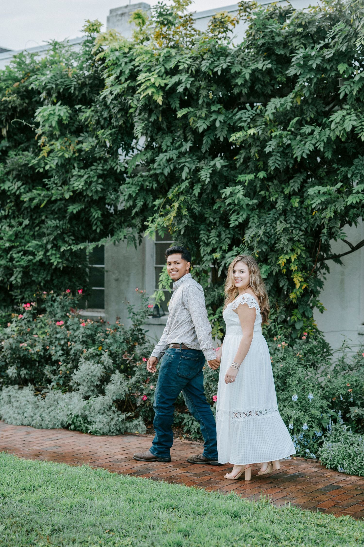 Two people walk through a serene garden pathway bordered by lush greenery and blooming flowers along a brick wall.