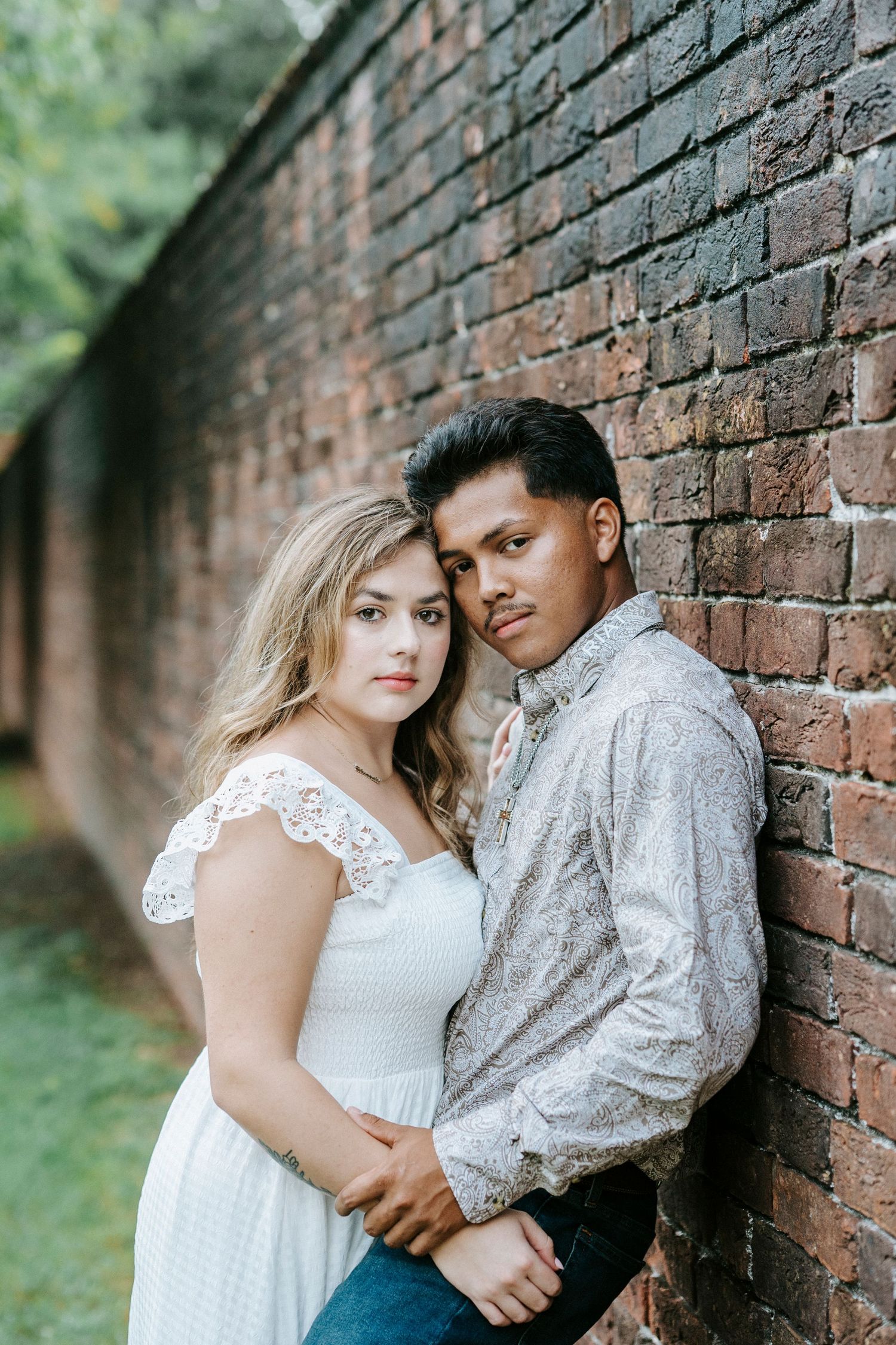 A couple poses intimately against a rustic brick wall in casual attire during an outdoor portrait session.