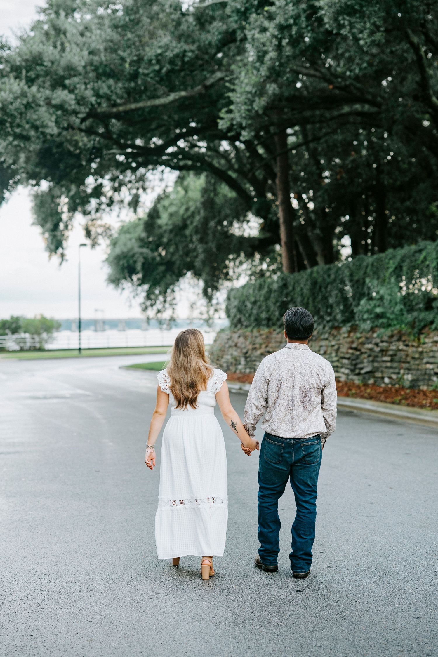 Couple walking hand in hand down a tree-lined street in casual attire, photographed from behind in a romantic setting.