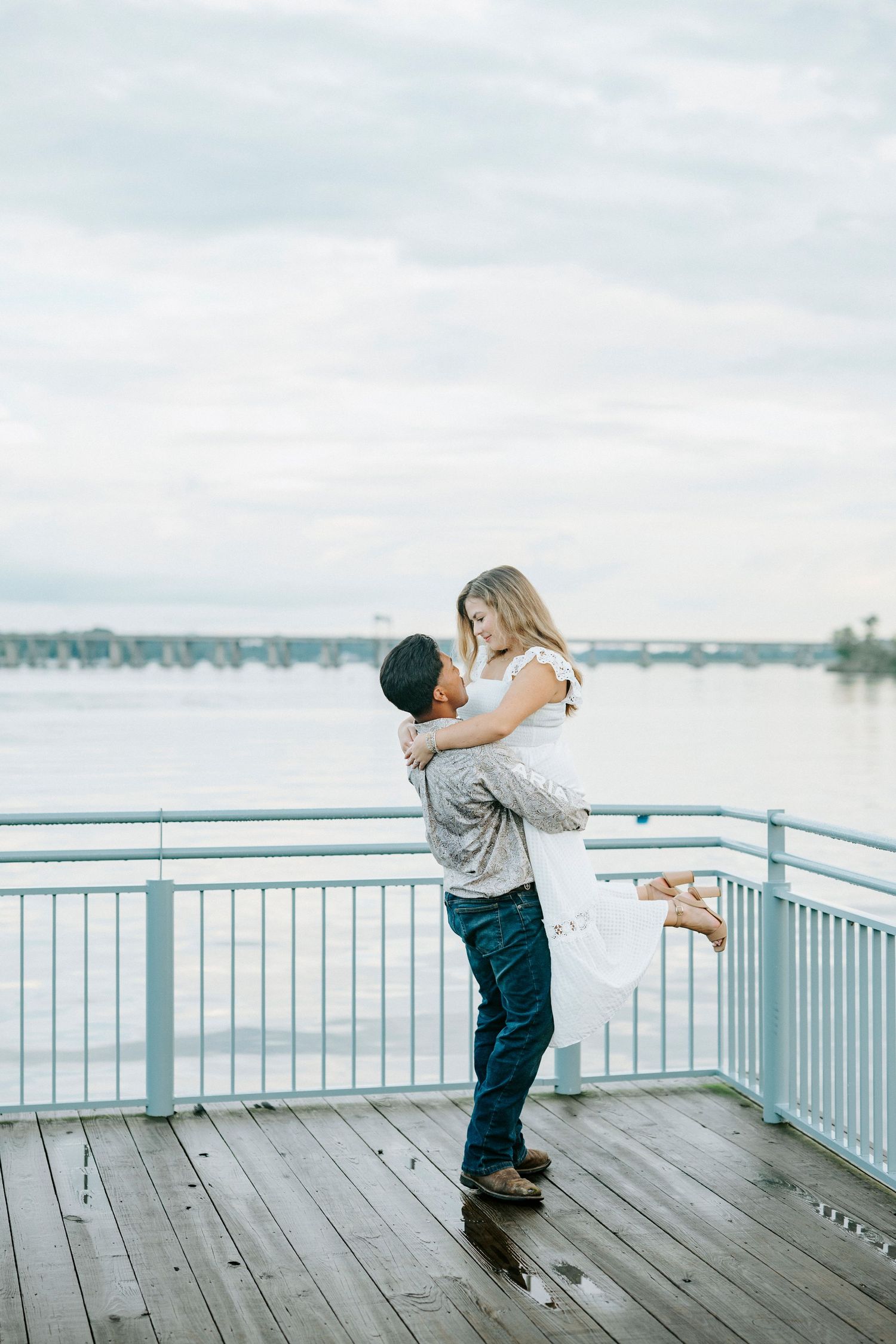 A couple twirls playfully together on a wooden pier overlooking a serene waterfront on a cloudy day.