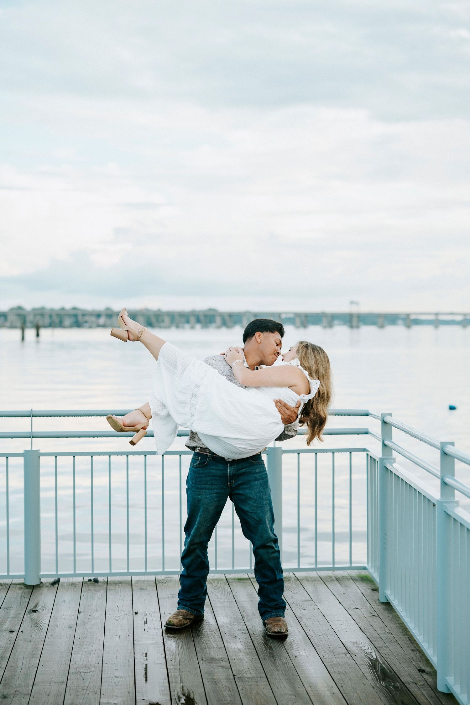 Two people in romantic poses on a waterfront deck overlooking a harbor during an engagement photoshoot.