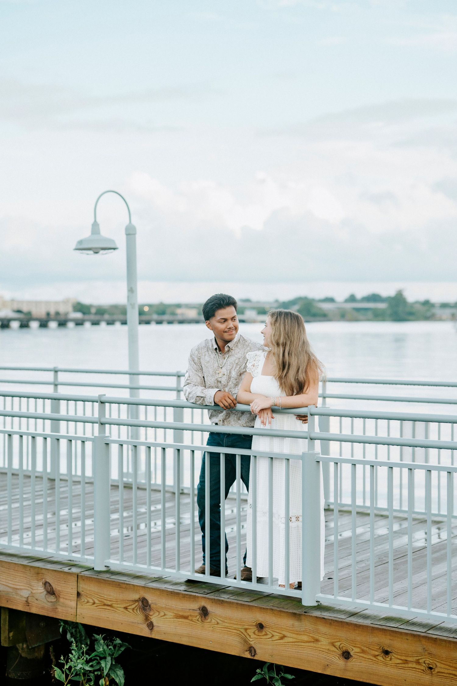 A romantic couple sharing a tender moment along a waterfront boardwalk with city views in the background.