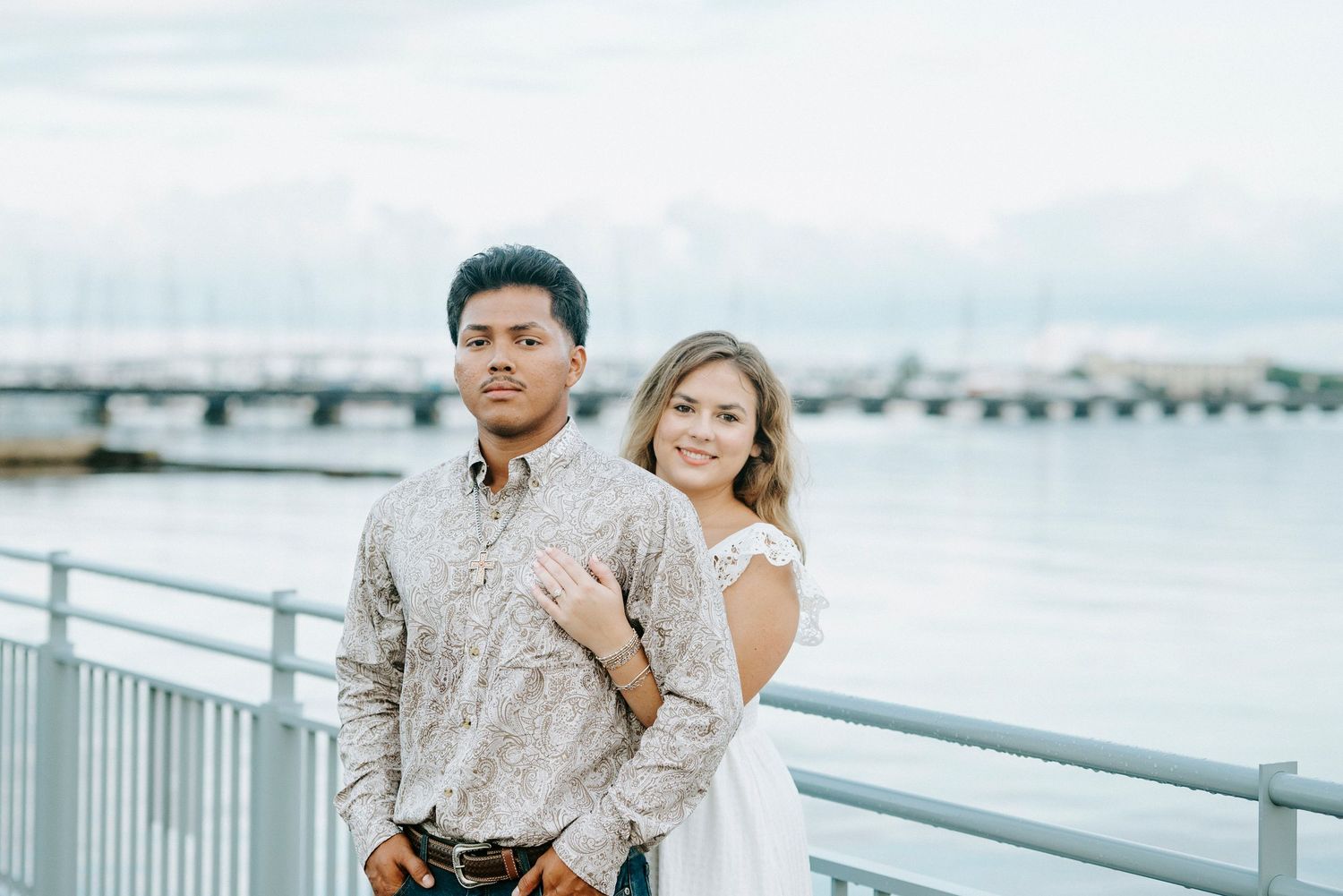 A couple shares a tender moment on a white pier overlooking the waterfront during a sunny day.