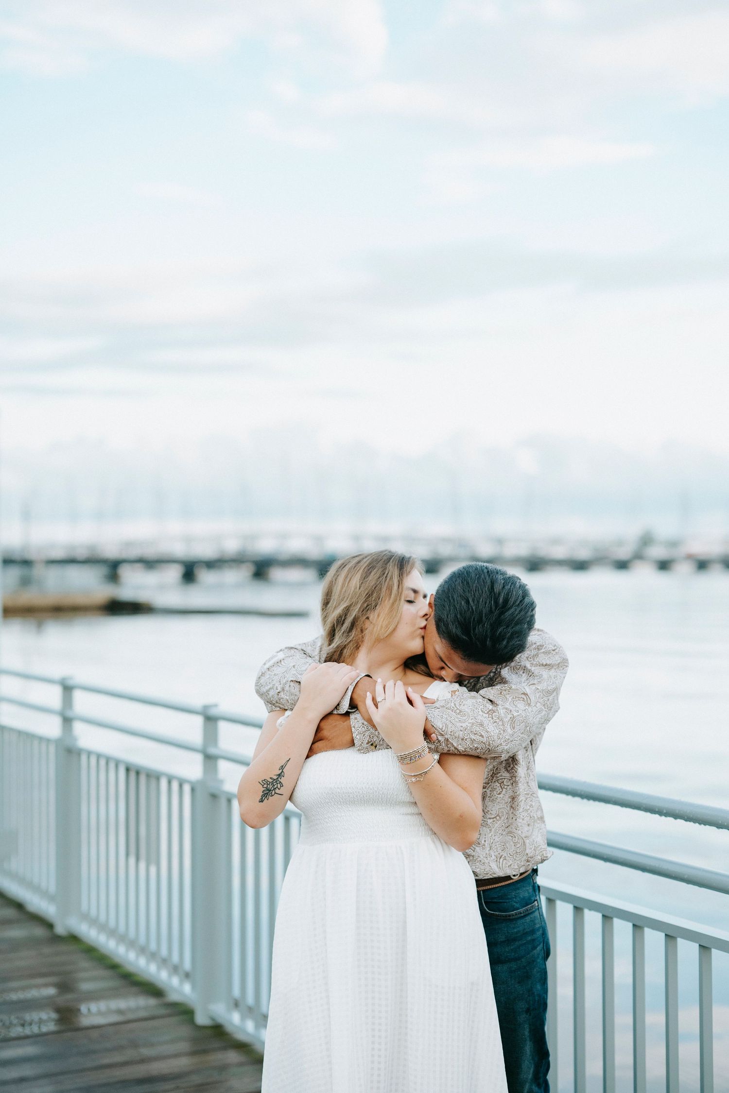 A romantic moment shared between a couple on a waterfront boardwalk with a bridge visible in the background.