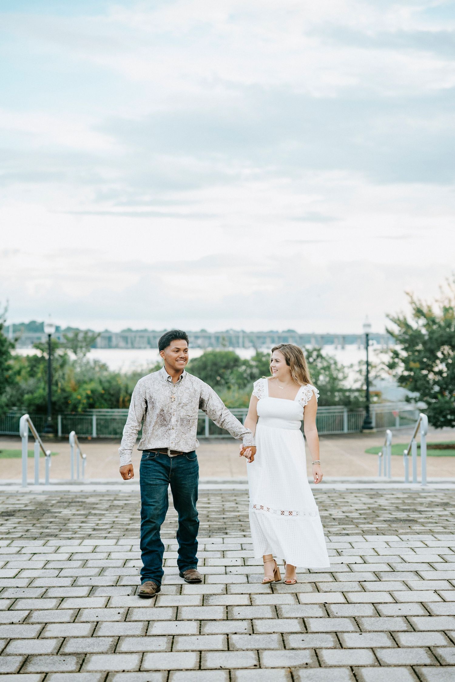 A couple dancing playfully on a stone paved plaza with waterfront views on a cloudy day.