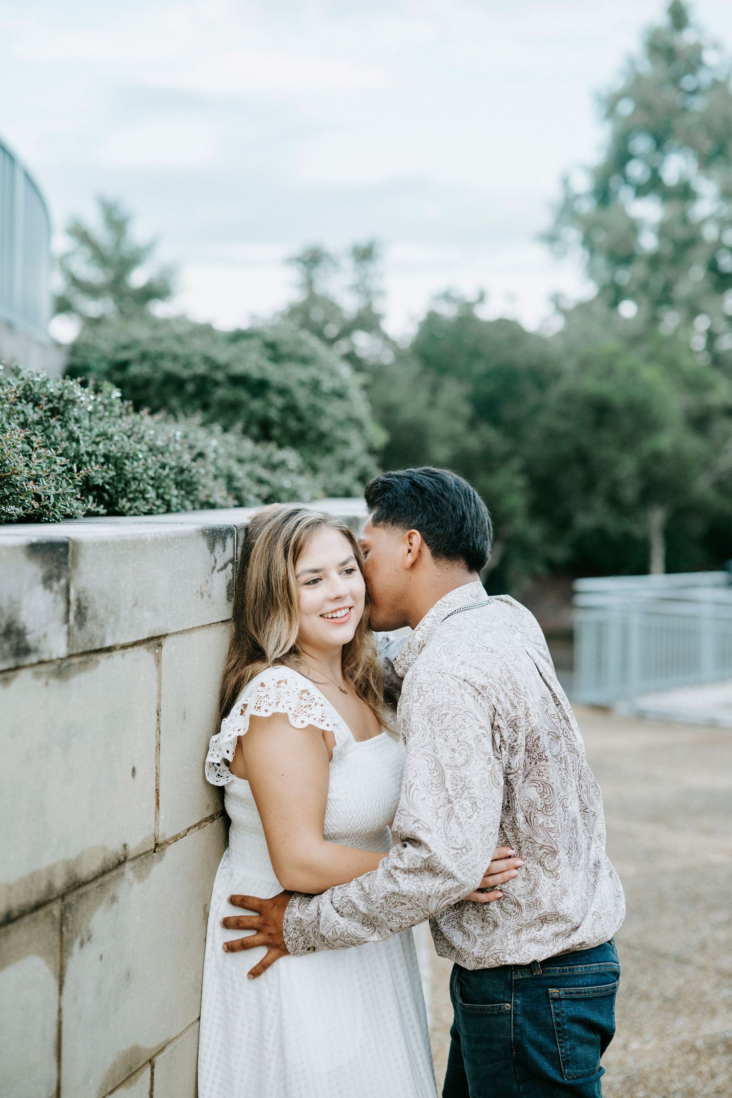 A tender romantic moment between a couple embracing outdoors against a stone wall with greenery in the background.