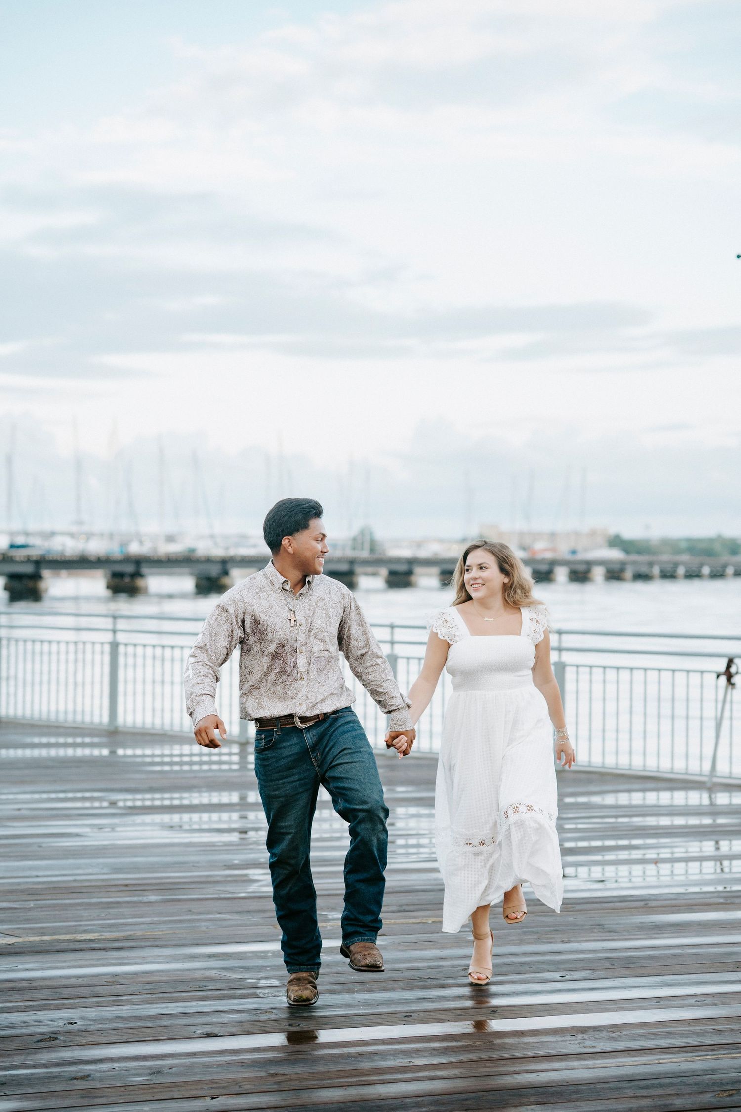 A couple walking and dancing together on a waterfront boardwalk with a bridge in the background.