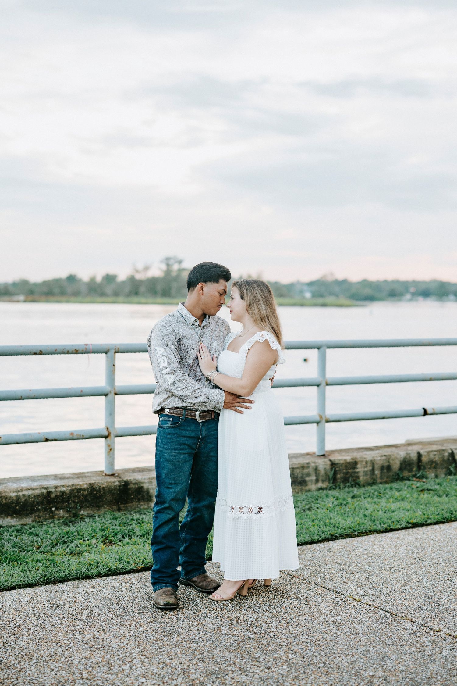 Two people share a romantic moment near a waterfront railing as they pose for engagement photos at sunset.