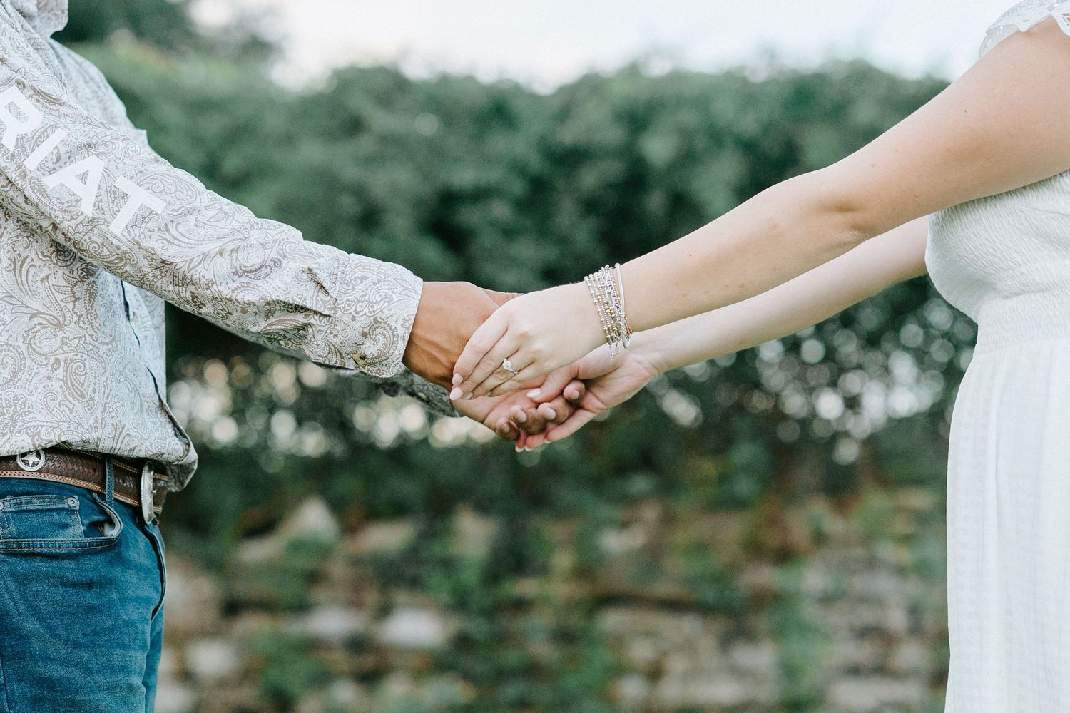 Close up shot of a bride and groom holding hands with wedding rings on their fingers outdoors.
