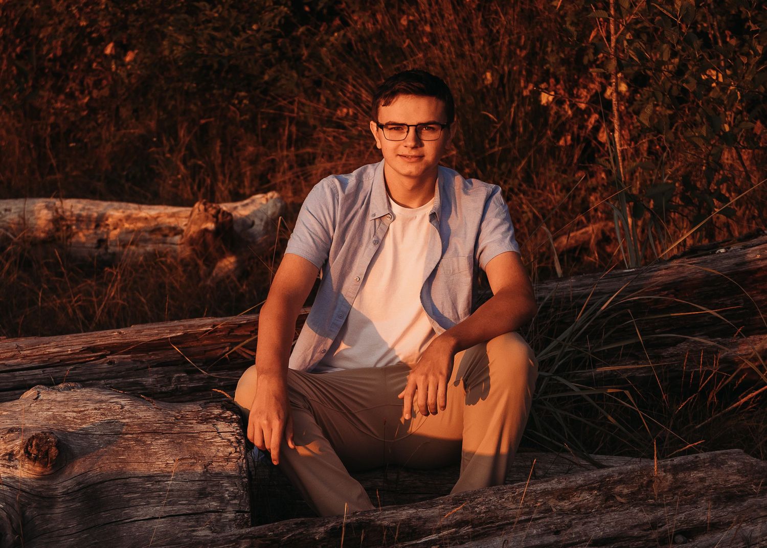A couple in casual attire embraces while sitting in tall grass during golden hour.