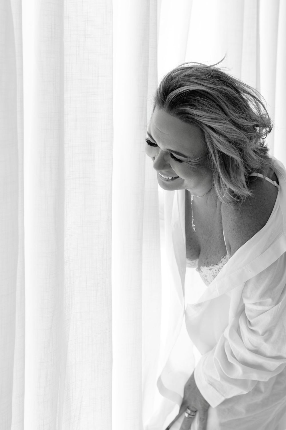 Black and white boudoir photo showcasing playful movement and natural light against white curtains.