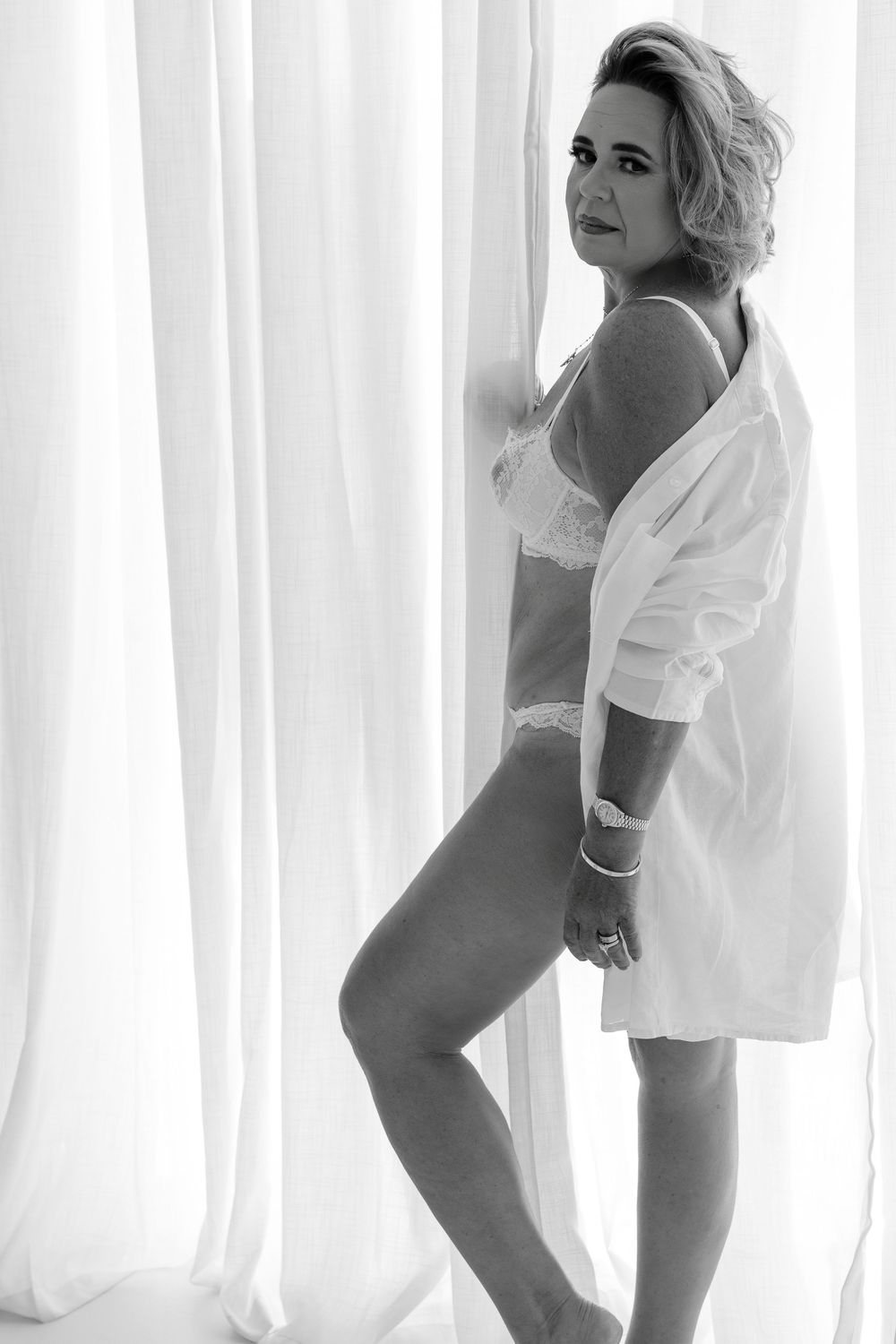 Monochrome intimate portrait showing a figure in lingerie and white shirt by sheer curtains.