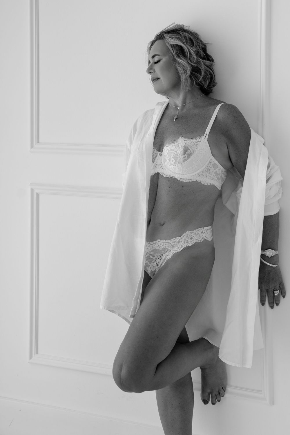 Monochromatic boudoir photo of a figure in white lace lingerie against a light backdrop.
