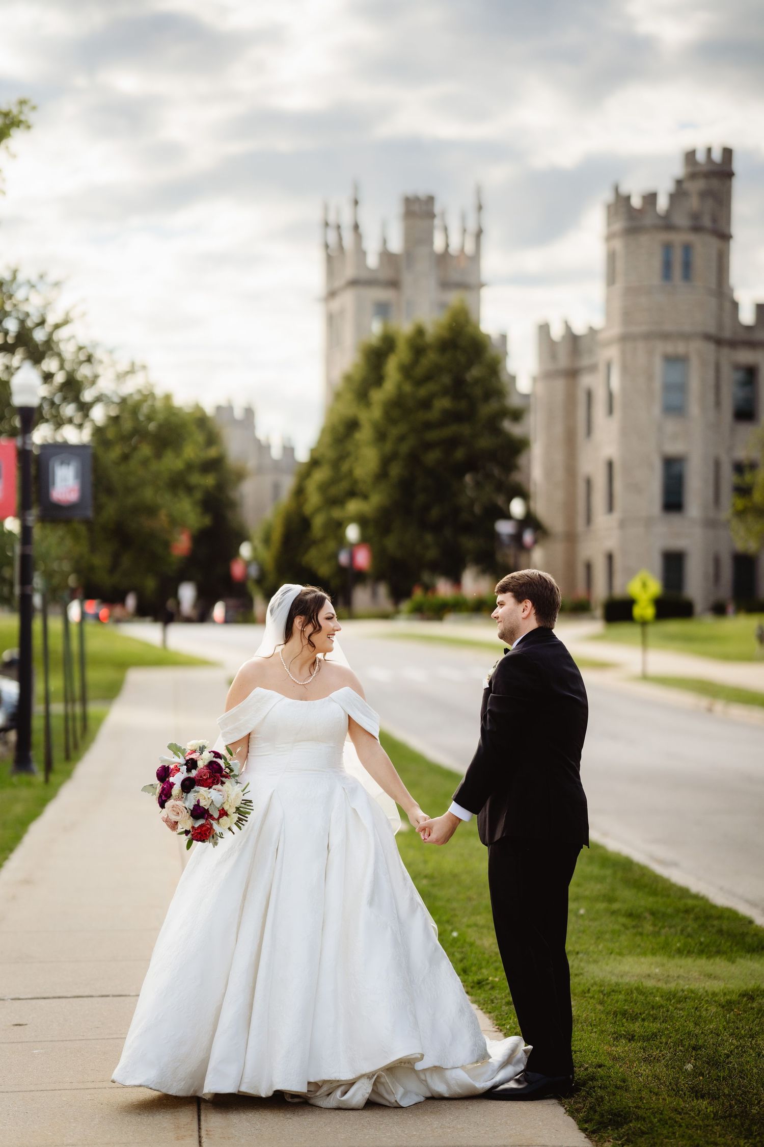 A couple in wedding attire poses on a path near a beautiful gothic-style castle with towers under cloudy skies.