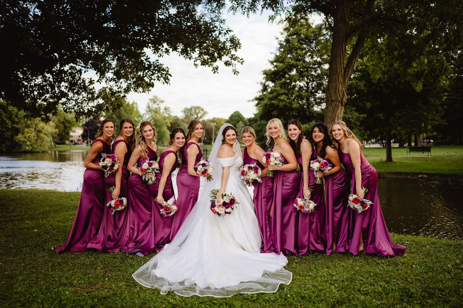Wedding party in burgundy bridesmaid dresses and white bridal gown pose together on grassy lawn near trees and lake.
