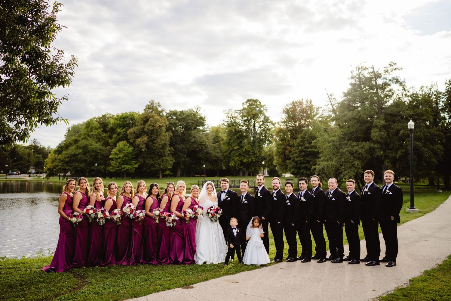 Wedding party in burgundy dresses and black suits poses outside on a lush garden path for formal portraits.