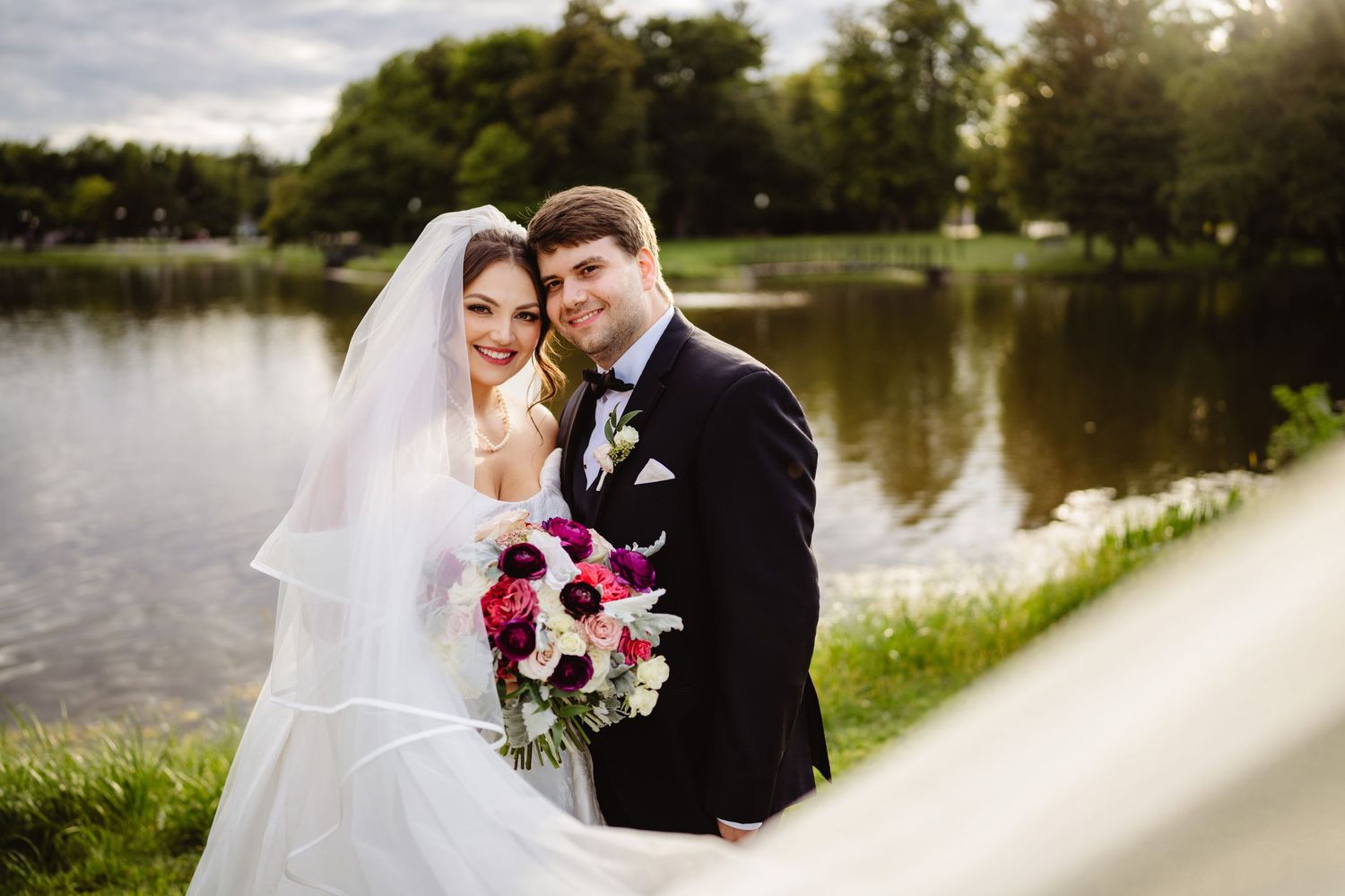 A romantic outdoor wedding photoshoot by a serene lake with trees and natural lighting in the background.