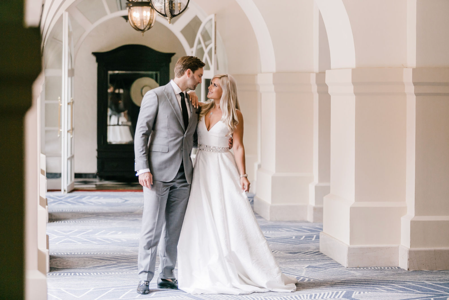 Wedding couple stands between classic white columns in an elegant architectural setting.