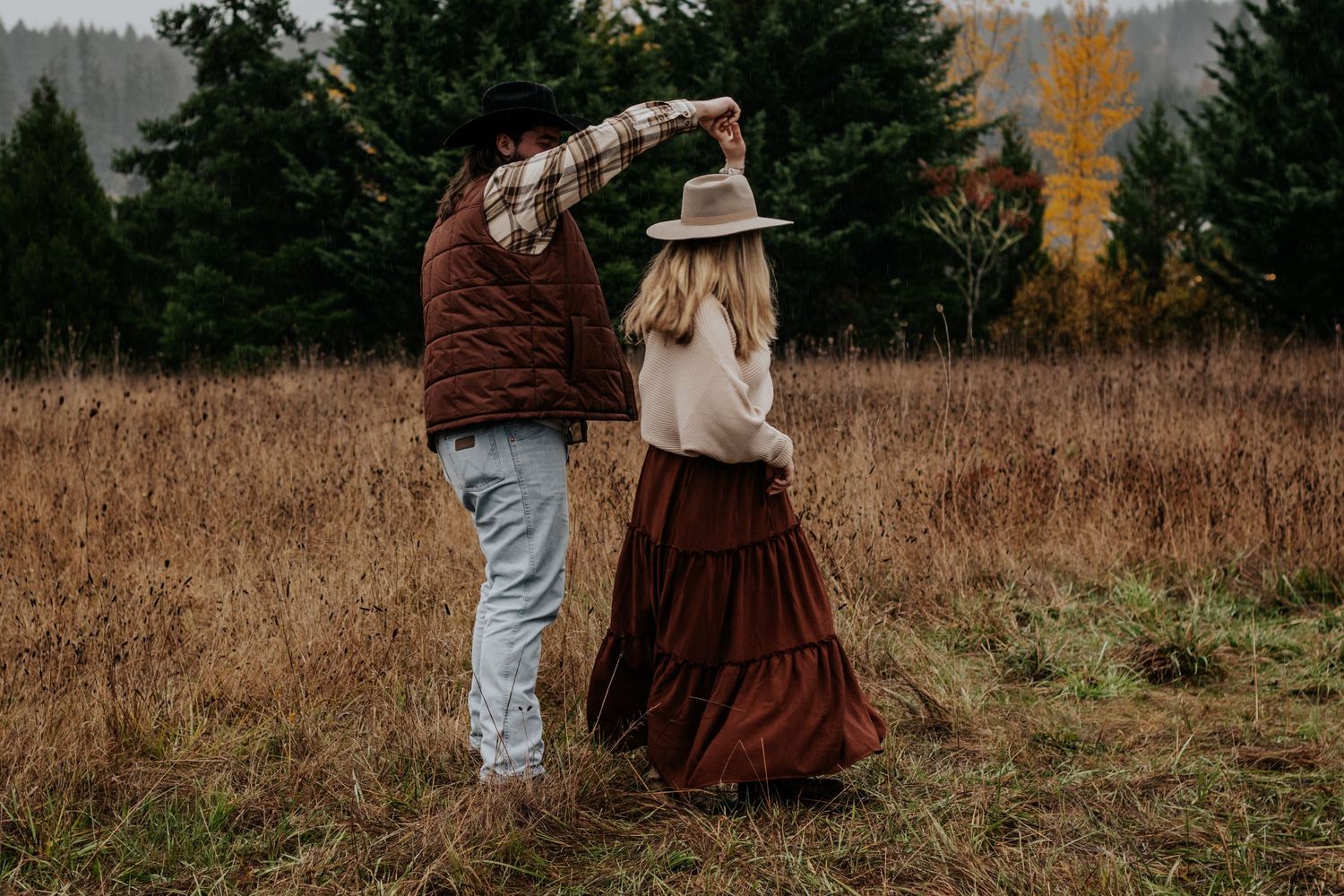 Couple dancing in autumn meadow surrounded by evergreens, woman in brown skirt and hat being twirled.
