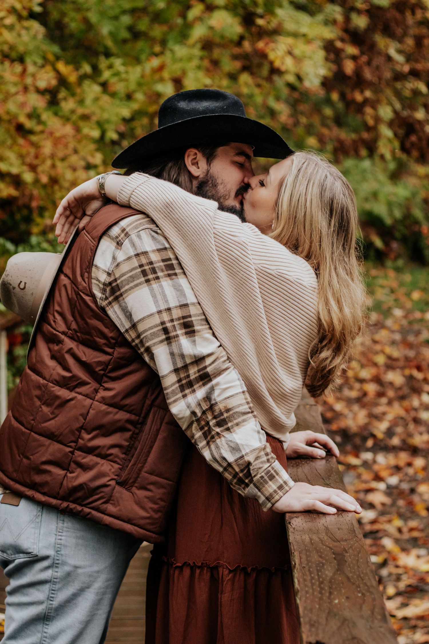 Couple sharing a kiss on autumn bridge, man in black cowboy hat and plaid shirt, woman in cream sweater.