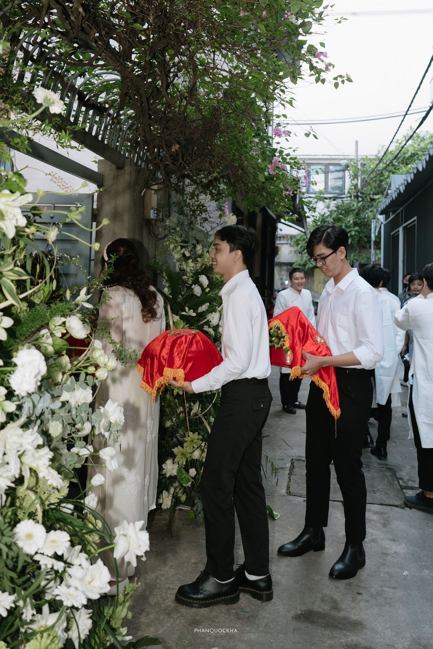 Two groomsmen in white shirts holding red ceremonial wedding gifts beside floral arch at Vietnamese wedding.