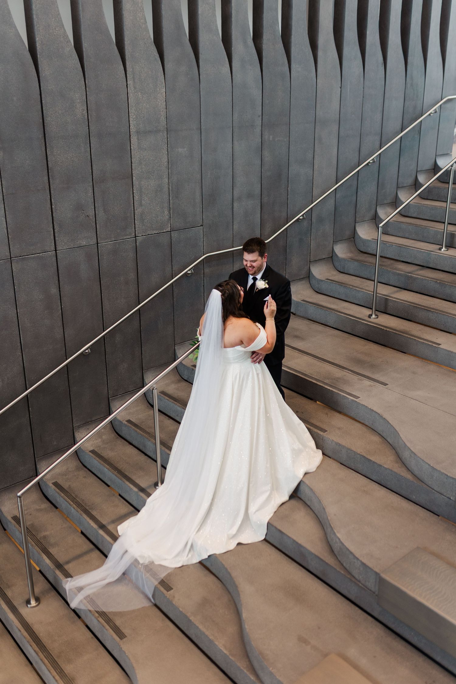 A couple in wedding attire embraces on modern concrete steps with a sleek grey architectural backdrop.