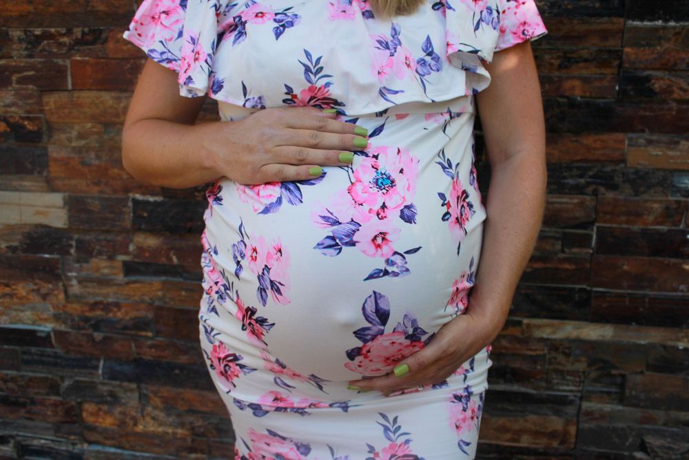 Pregnant belly shown in a white dress with pink and purple floral pattern against brick wall.