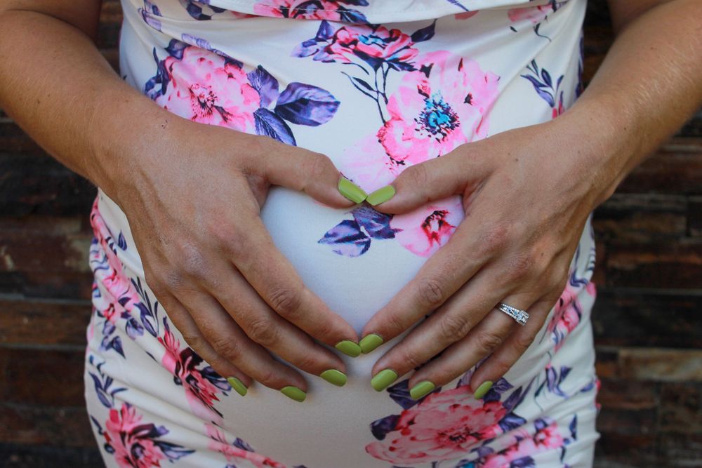 A pregnant woman in a floral pink dress cradles her baby bump while standing against a brick wall background.