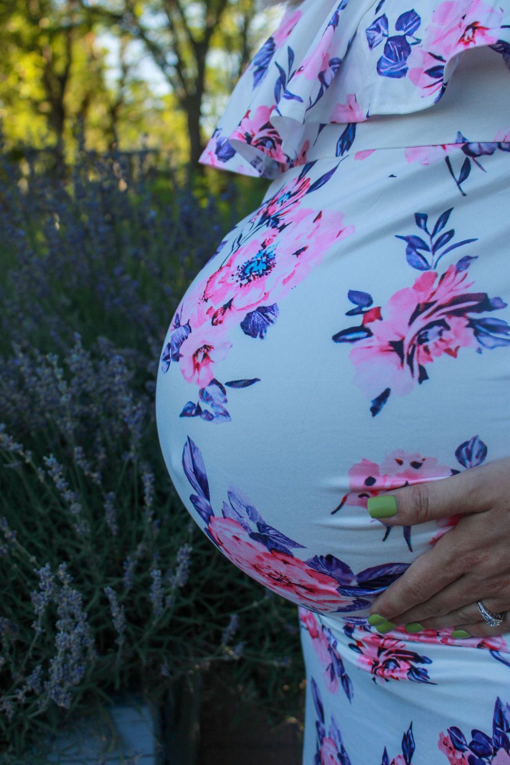 Side view of a pregnant belly covered in a white dress with pink and blue floral print against a garden backdrop.