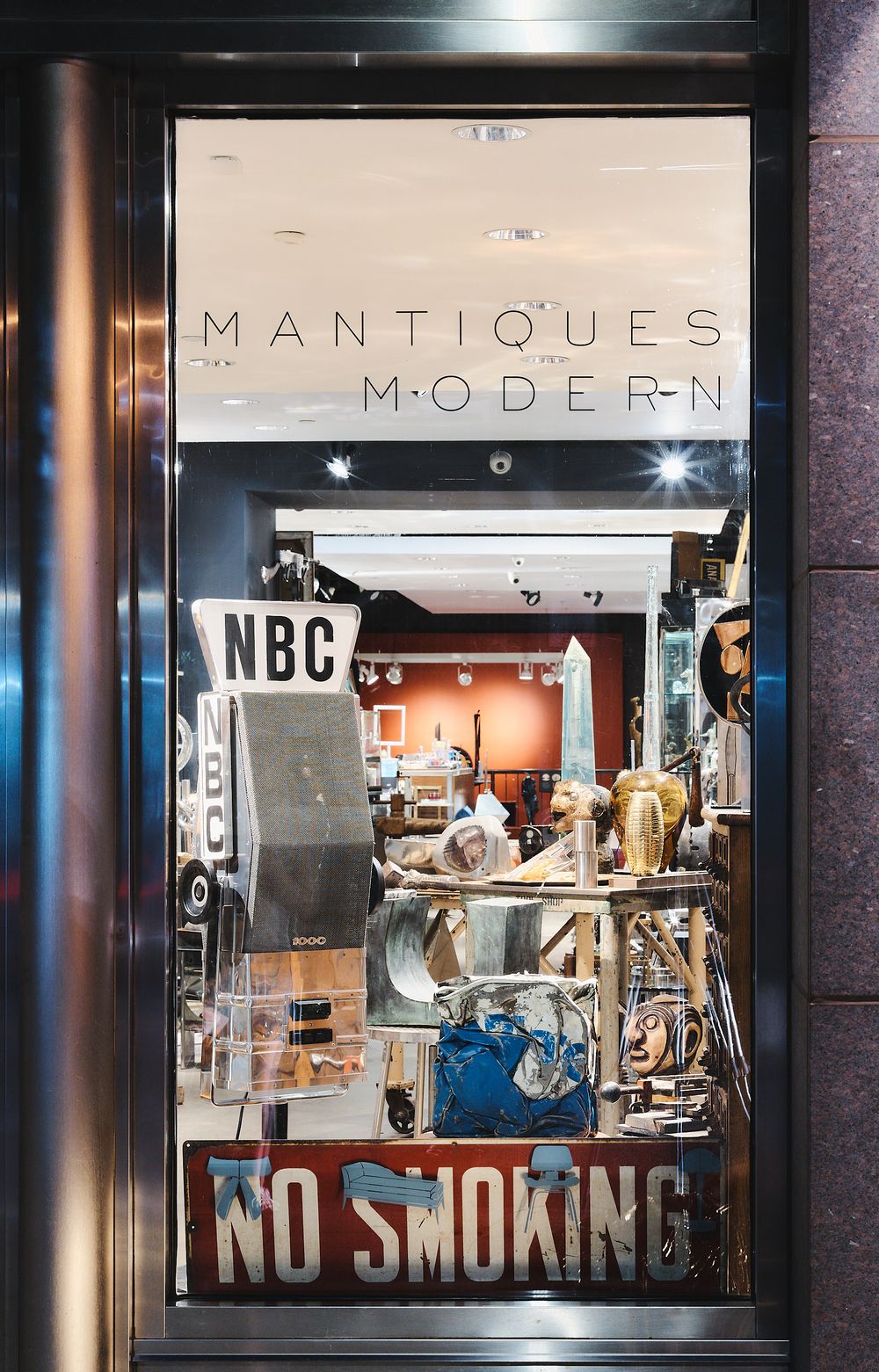 A well-lit storefront window display shows modern design elements with mannequins and a 'No Smoking' sign at night.