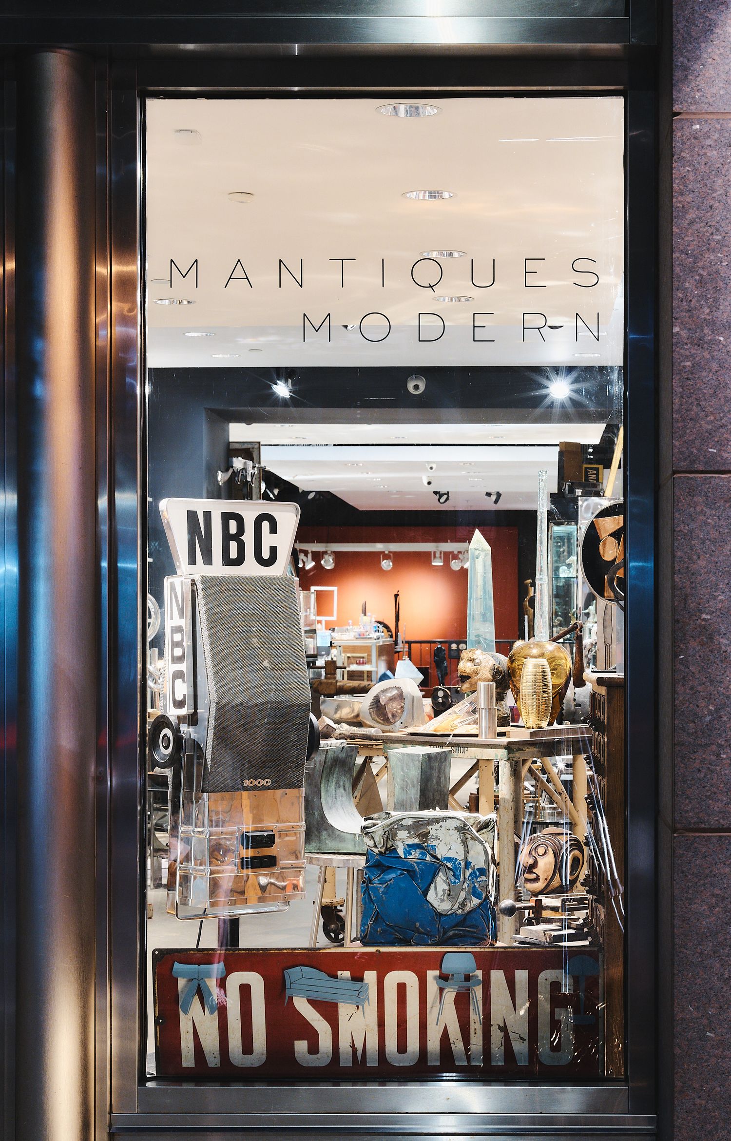 A well-lit storefront window display shows modern design elements with mannequins and a 'No Smoking' sign at night.