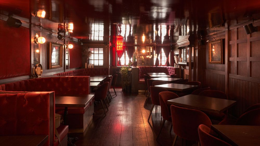 A dimly lit rustic restaurant interior with wooden beams, red lighting, and empty dining tables creating a cozy ambiance.