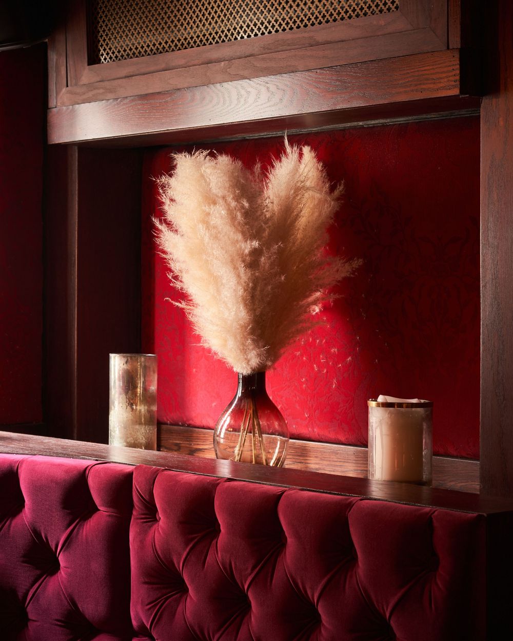 A white pampas grass plume in a vase sits against a red wall with plush burgundy velvet seating below.