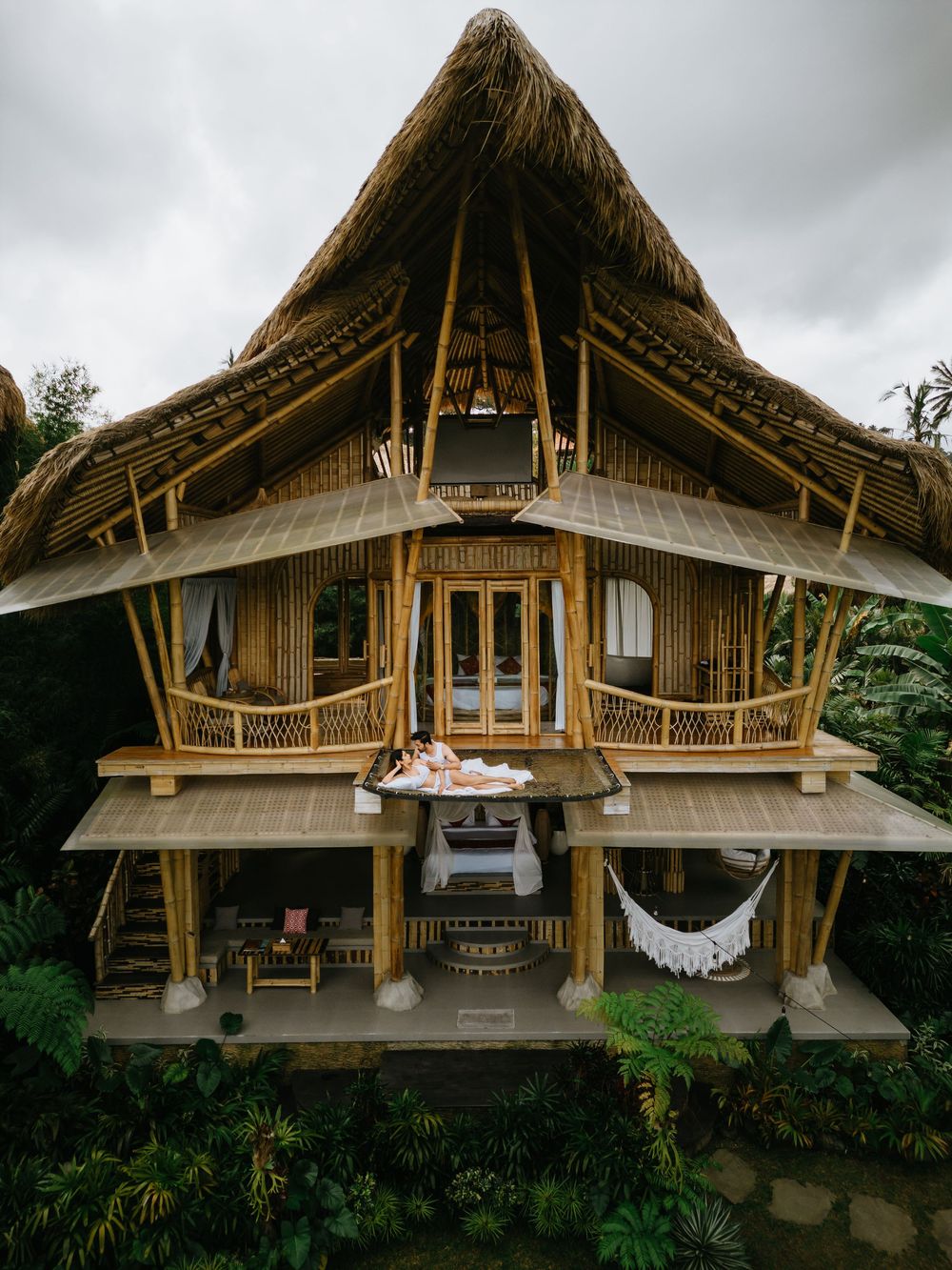 A luxurious two-story bamboo villa with thatched roof and hammock nestled in tropical forest.