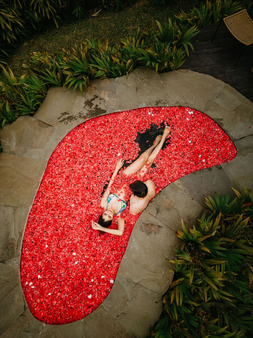 Aerial view of a person lounging on a red bean-shaped pool float surrounded by tropical plants.