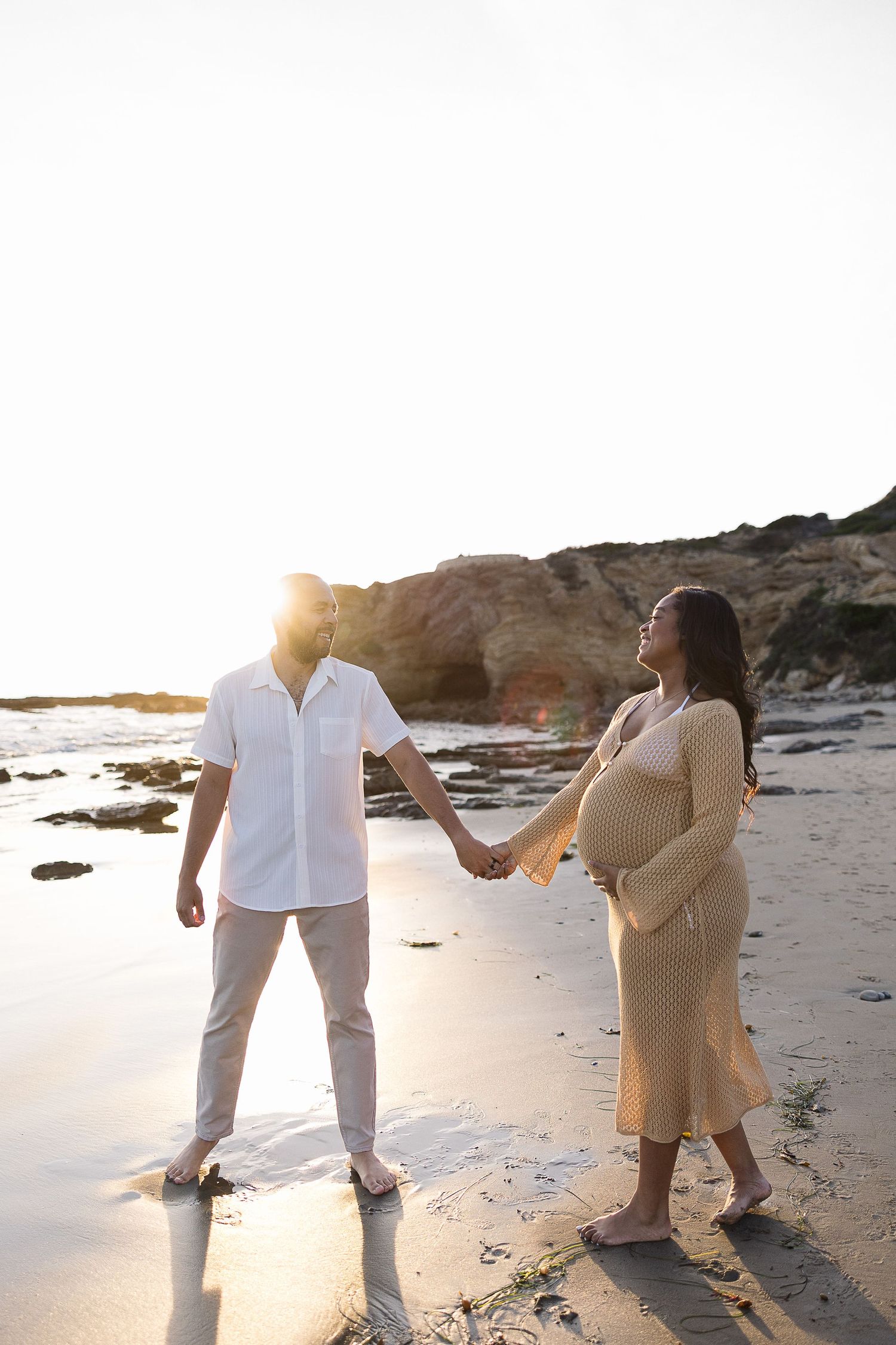 Expectant couple holding hands on beach at sunset with rocky cliffs and tide pools in background.