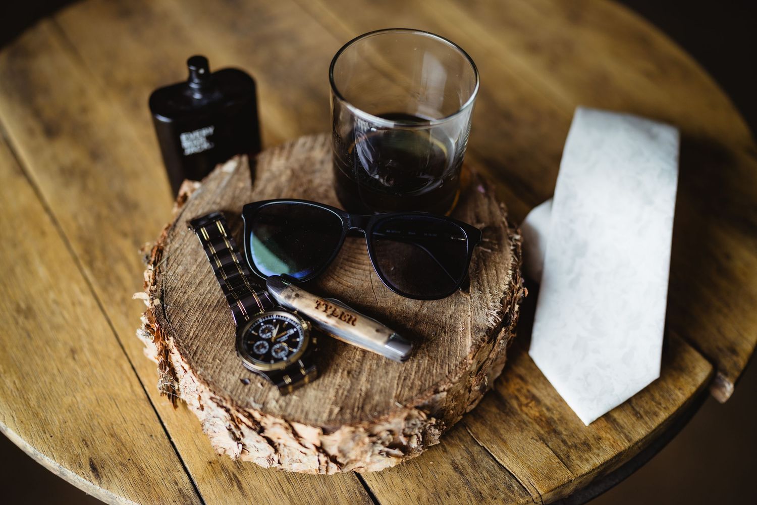 Sunglasses and drinking glass arranged on rustic wood slice serving board with dark bottles on wooden table.