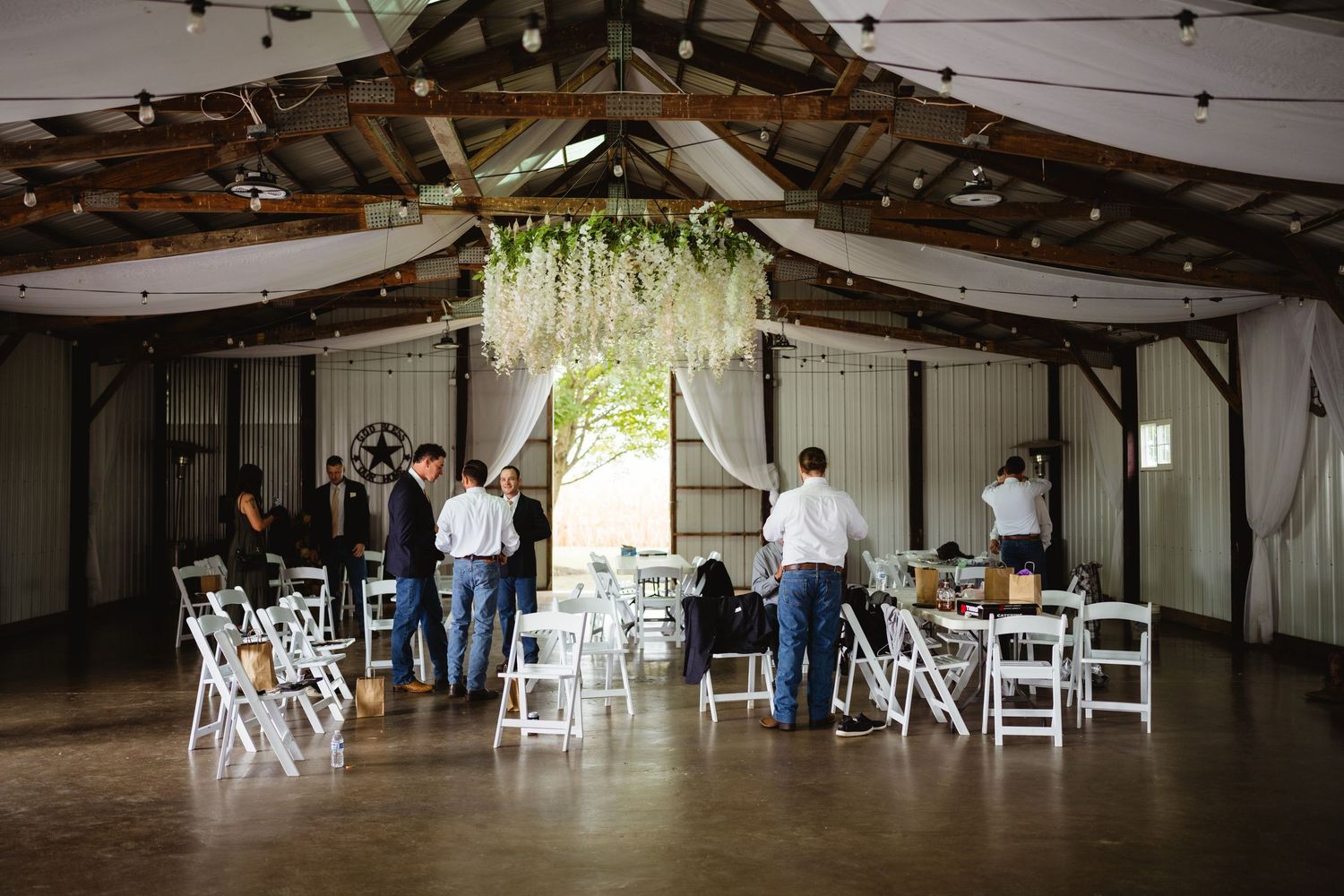 Rustic barn venue being set up for a wedding ceremony with white chairs and hanging floral decor.