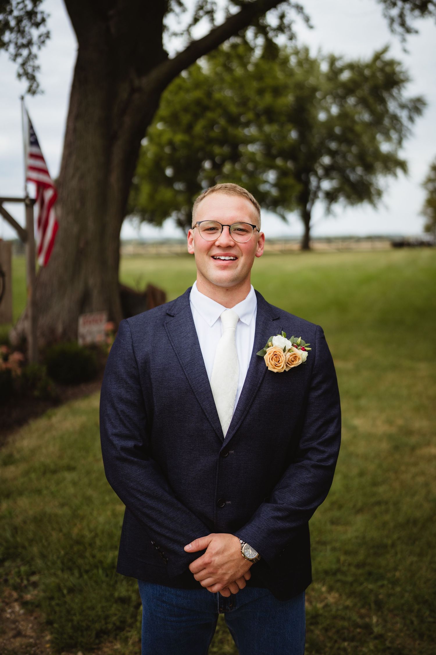 Groomsman poses outdoors wearing a dark navy suit with a floral boutonniere against a tree-lined background.