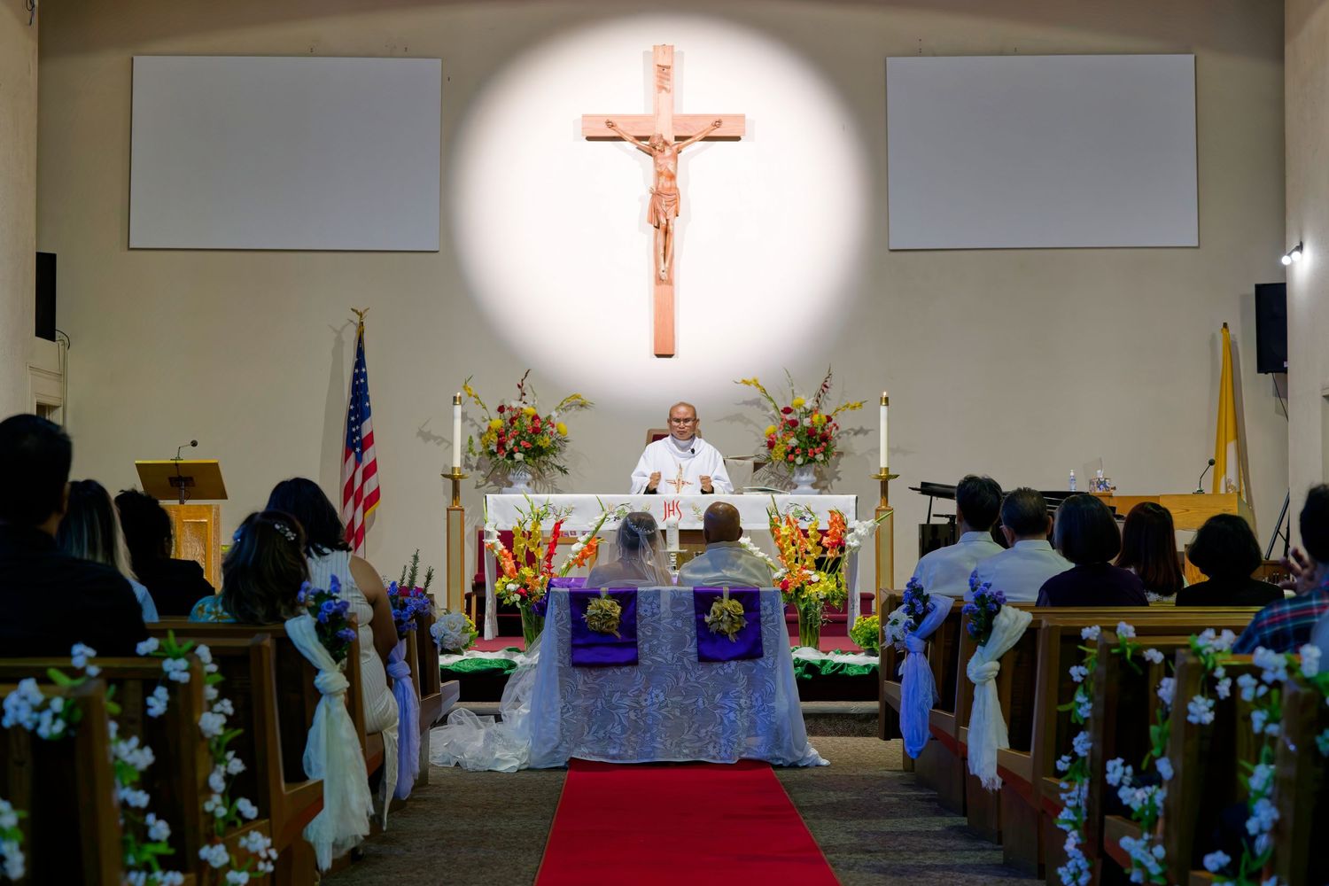 A church ceremony setup with multiple pews, an illuminated crucifix, and floral decorations at the altar.