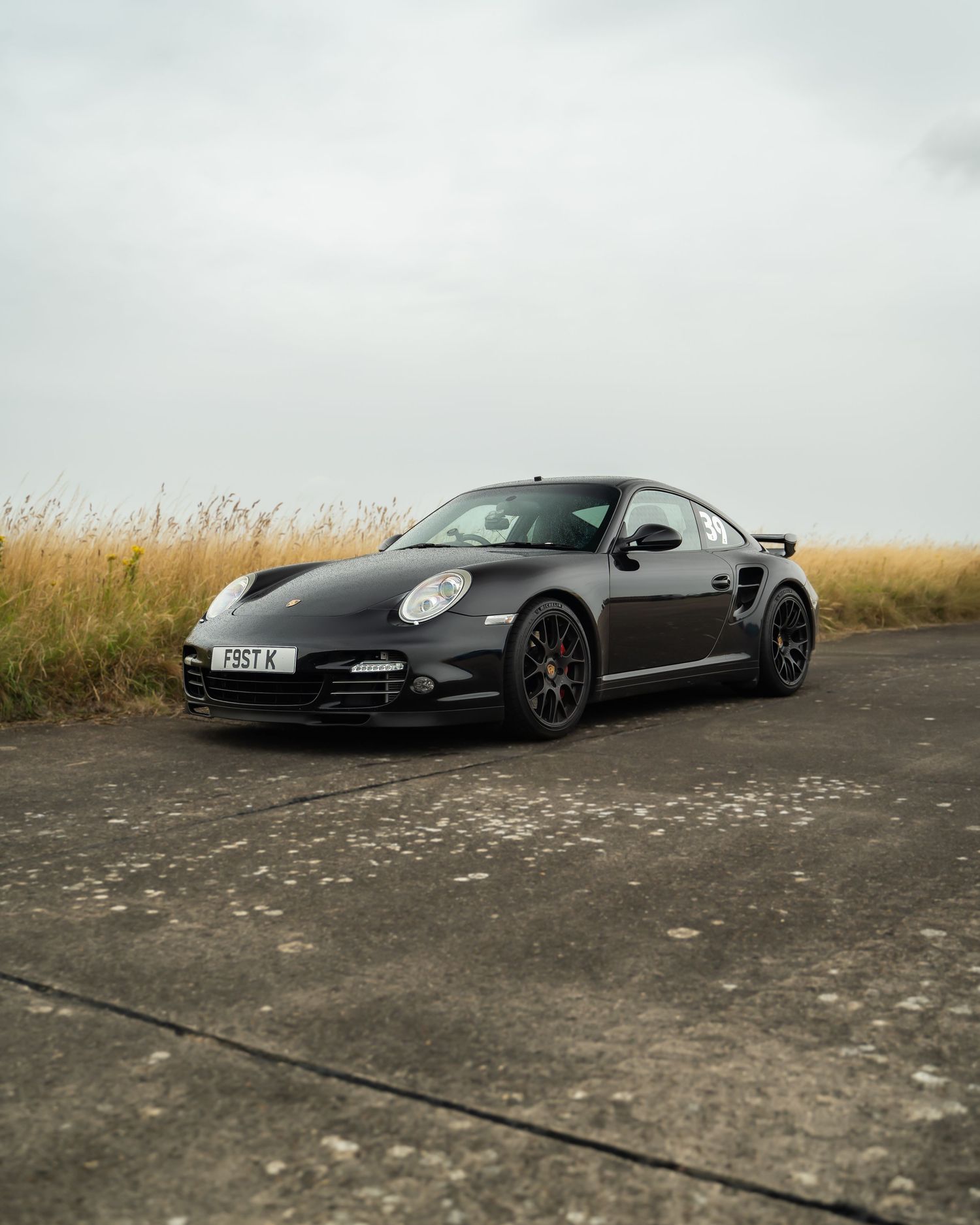 Black Porsche 911 sports car parked on concrete against cloudy sky.