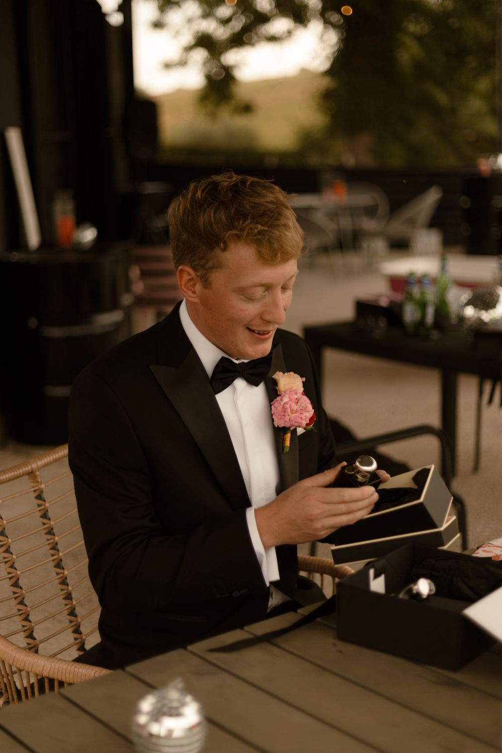 A formally dressed guest checks their phone while seated at an outdoor wedding reception table at dusk.