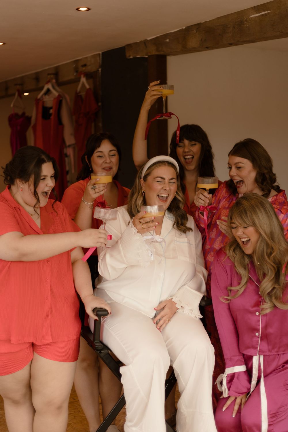 A group wearing pink and coral outfits laughing and celebrating together in a bridal preparation room.