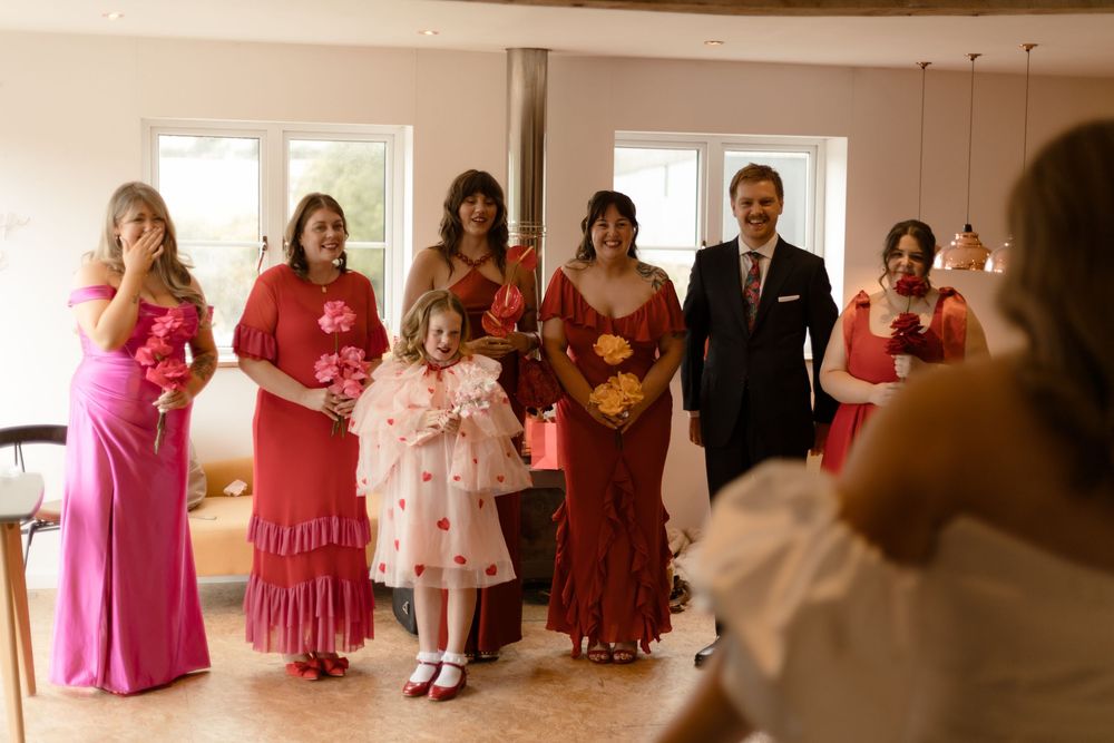 A lineup of people in pink and red formal attire walk through an indoor venue in sequence.