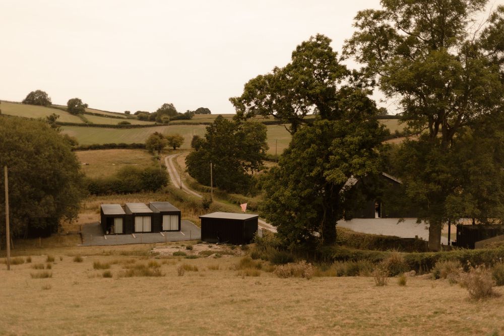 Rustic countryside landscape with rolling hills, scattered trees and an old stone building in warm sepia tones.