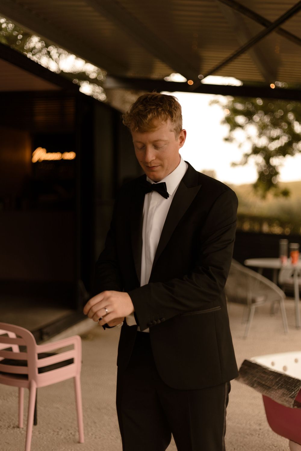 A person in formal black tuxedo attire stands outdoors at sunset in a candid moment.