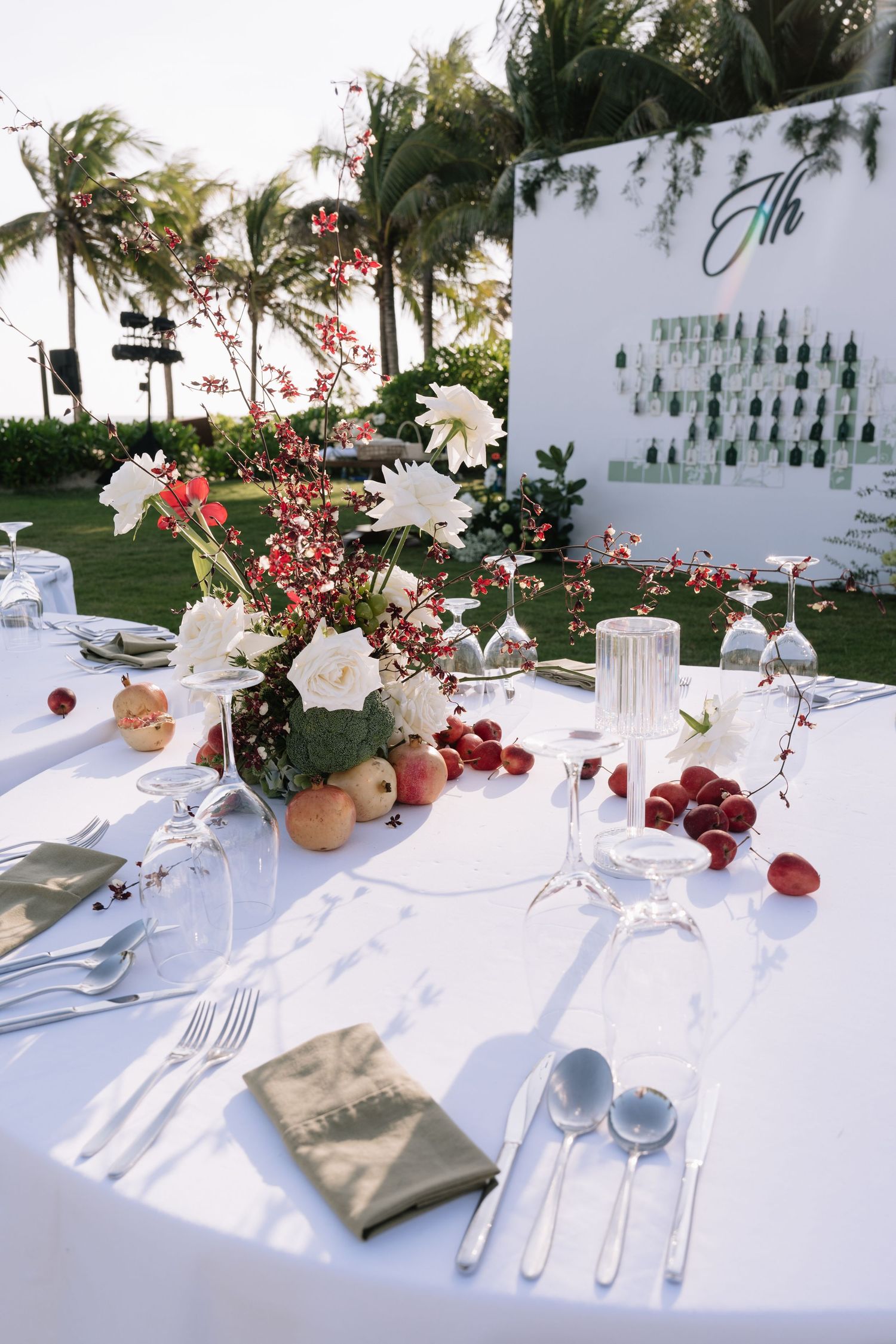 Tropical outdoor wedding reception table with white florals, red berries, fruits, and glassware near palm trees.