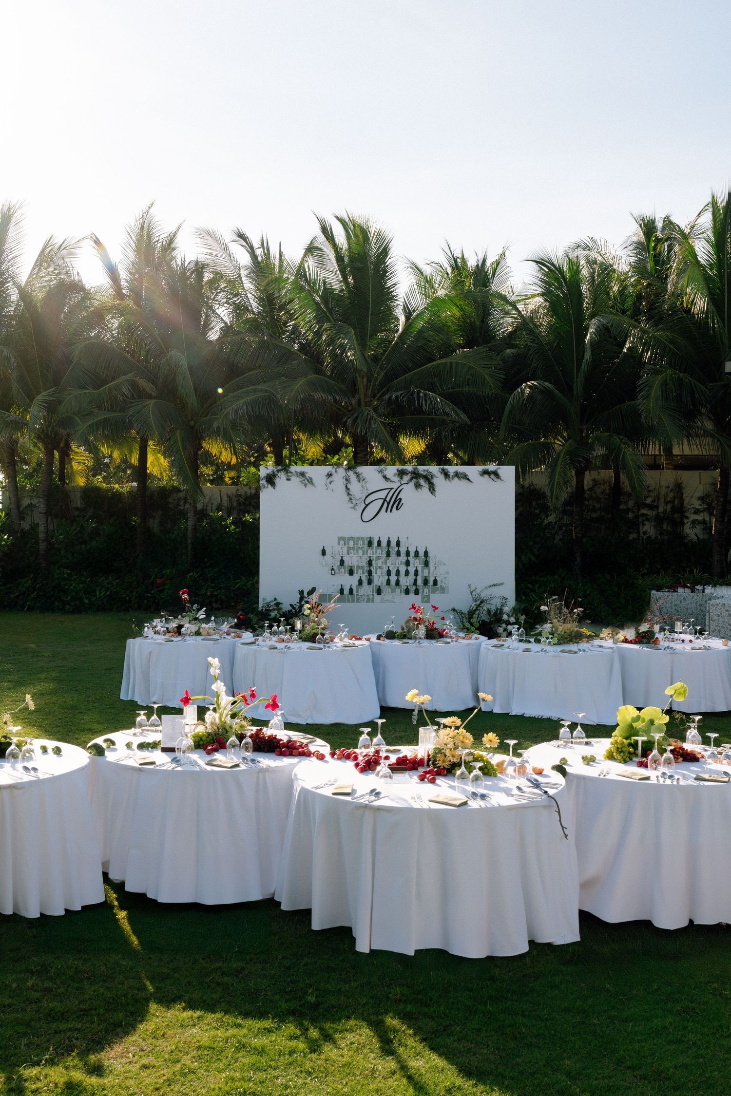 Outdoor garden wedding reception with round white-draped tables and a decorative monogram backdrop amid palm trees.