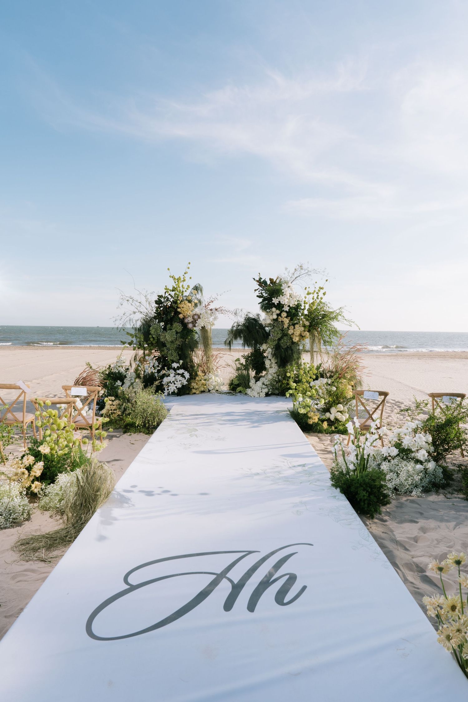 White aisle runner with monogram leading to a floral arch wedding ceremony setup on a sandy beach.